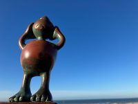 A small bronze sculpture depicting a human-like figure with a big round belly, big feet and a sad face, standing with hands to its head against a clear blue sky.